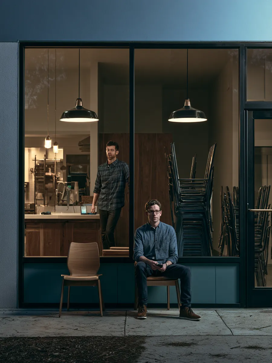Wide dusk view of a small cafe storefront as two staff members stack chairs and dim the pendant lights