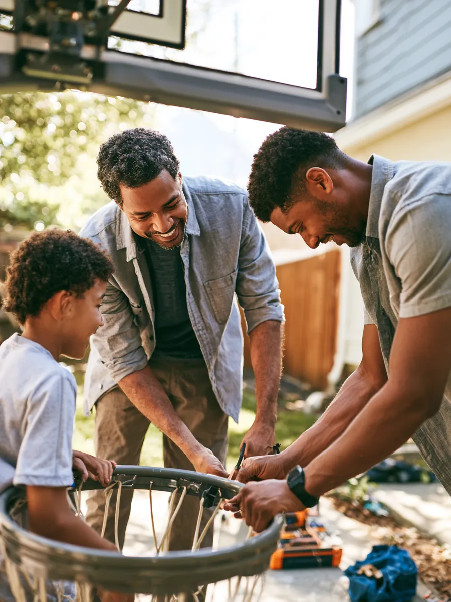 Father and son assembling a new basketball hoop at the edge of a backyard driveway